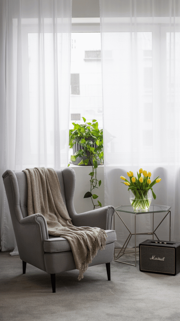 A photograph of a serene, minimalist living room bathed in soft, natural light. A comfortable gray upholstered armchair with a textured beige throw blanket sits on a light gray carpet, gently angled towards the window. Through sheer white curtains, a vibrant green potted plant with trailing vines cascades from a windowsill, adding a touch of life to the room. A geometric metal side table beside the chair holds a clear glass vase filled with bright yellow tulips, while a black Marshall speaker rests on the floor, completing the clean, modern aesthetic.